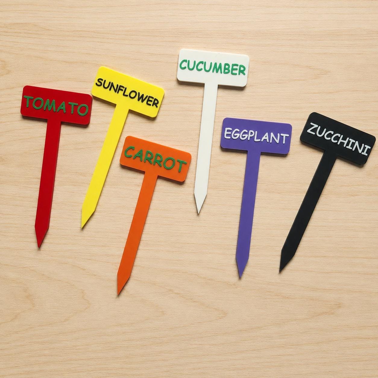 A set of six colorful garden plant markers in red, yellow, orange, white, purple, and black. Each T-shaped stake features raised lettering identifying the plants as TOMATO, SUNFLOWER, CARROT, CUCUMBER, EGGPLANT, and ZUCCHINI respectively.