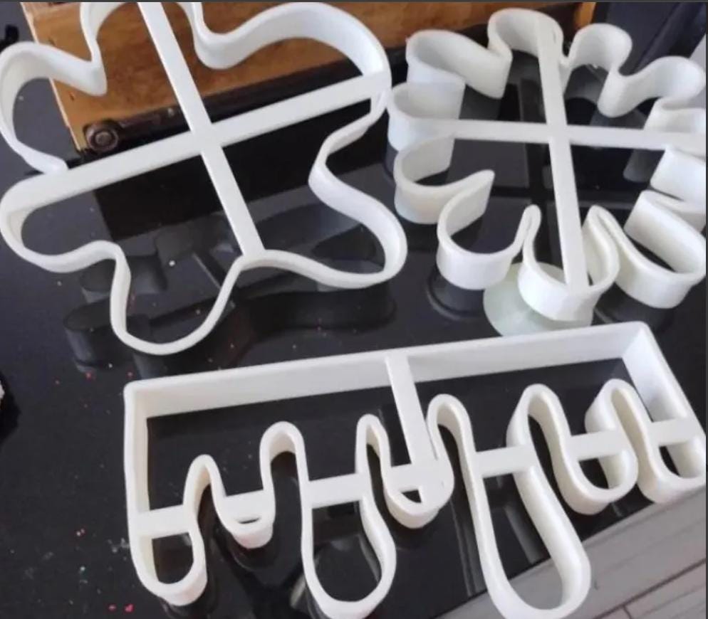 Three white plastic cookie cutters in various floral and abstract shapes. The cutters are designed with curved edges and cross-shaped supports, ideal for baking. The cutters are on a reflective black surface.