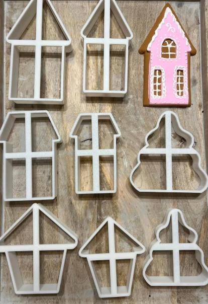 A collection of white plastic house-shaped cookie cutters and a decorated gingerbread house on a wooden surface. The gingerbread house is pink with white icing details.